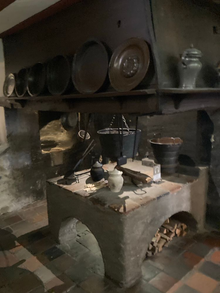 Eine historische Küche mit einer großen Kochstelle in der Mitte. Auf dem Herd Töpfe und Gefäße. Am Kamin lehnen große Zinnteller und Kannen. 