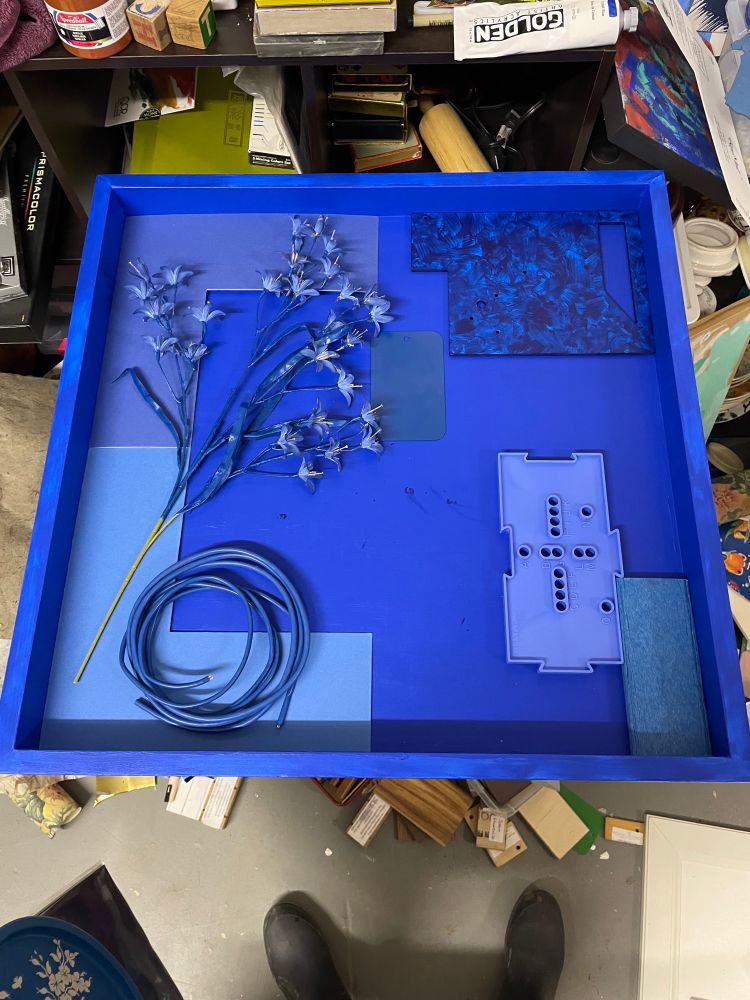 A blue shadow box frame with various shades of blue things in it (mat corners, random plastic, faux flowers, MDF or Masonite scrap, cable). My messy studio floor and disorganized shelf can be seen around it.