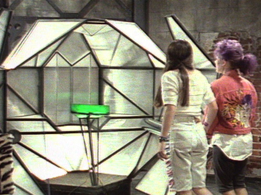 A still from The Girl From Tomorrow, an Australian young people's TV show from the 90s.

In the picture we see Alana and Jenny standing in front of the Time Capsule.

It has a long description in Wikipedia, the first part follows:

The Time Capsule is a time exploration vehicle. It was invented by a scientist named Bruno in the year 3000. The Capsule is exactly 1.7 m (5 ft 7 in) in height and width. Its composition in the series is unspecified, but as a real prop, it is a dark green steel frame model of a Rhombicuboctahedron plated with semidiagonal steel bars and plated with semitransparent plexiglas from the inside, except on the front side which is completely transparent.

More at https://en.wikipedia.org/wiki/The_Girl_from_Tomorrow#The_Time_Capsule