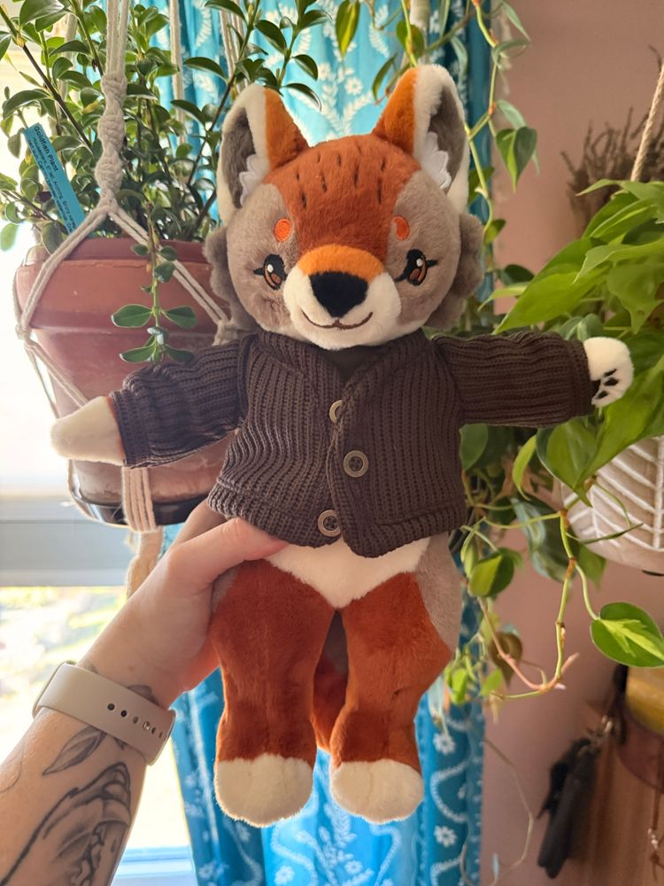 A coyote doll like plush dressed in a dark brown cardigan with buttons is held in front of a pink wall, teal patterned curtain and green plants. 