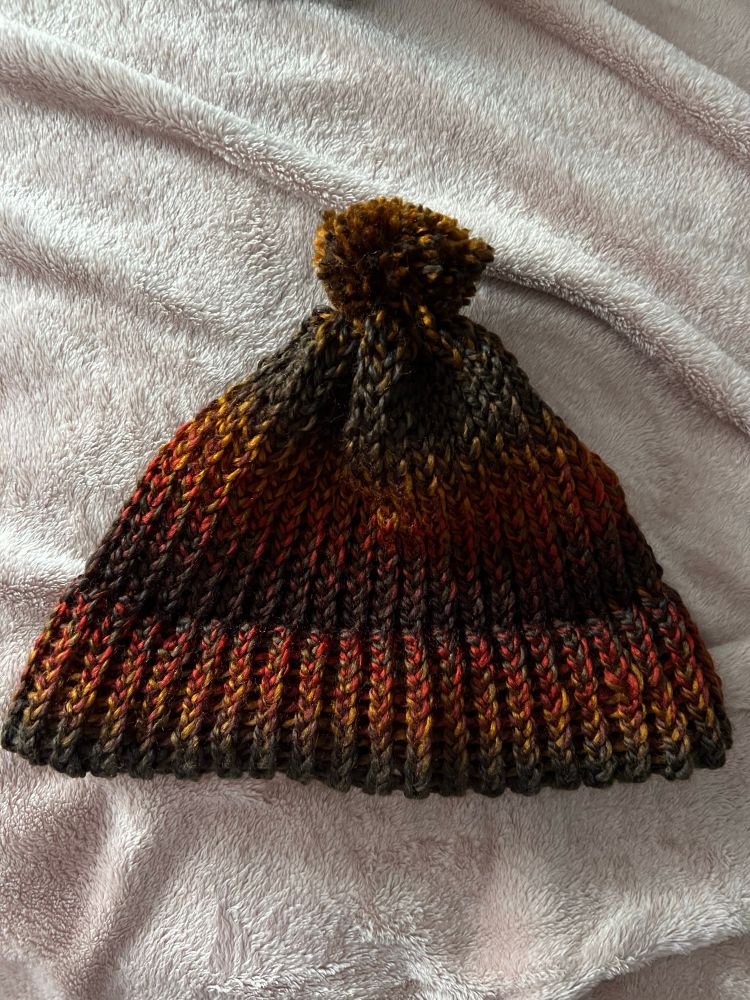 A loom knit acrylic toque/beanie in gradient deep reds, yellows, and greens. There is a Pom Pom in matching yarn.