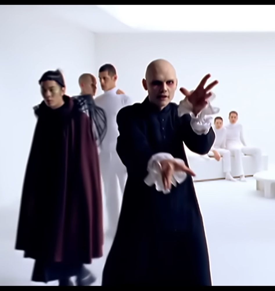 A frame of the Ava Adore music video with Billy Corgan in the foreground with frilly cuffs and James Iha and some actors in background.