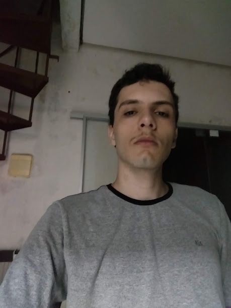 Homem de pele clara, olhar "caído" em direção à câmera, cabelos pretos e bigode fino. Usa uma blusa cinza. Está em um ambiente interno; há uma porta de vidro a suas costas e um pedaço de escada circular do outro lado, no canto superior.