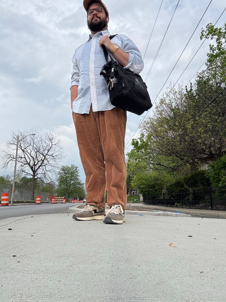 A 32 year old man waiting for the bus in a button up shirt and corduroy pants with a work bag