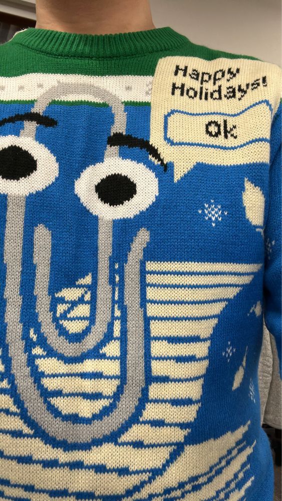 A Microsoft Clippy holiday sweater. Clippy is atop a stack of papers, with a popup that reads "Happy Holidays!"