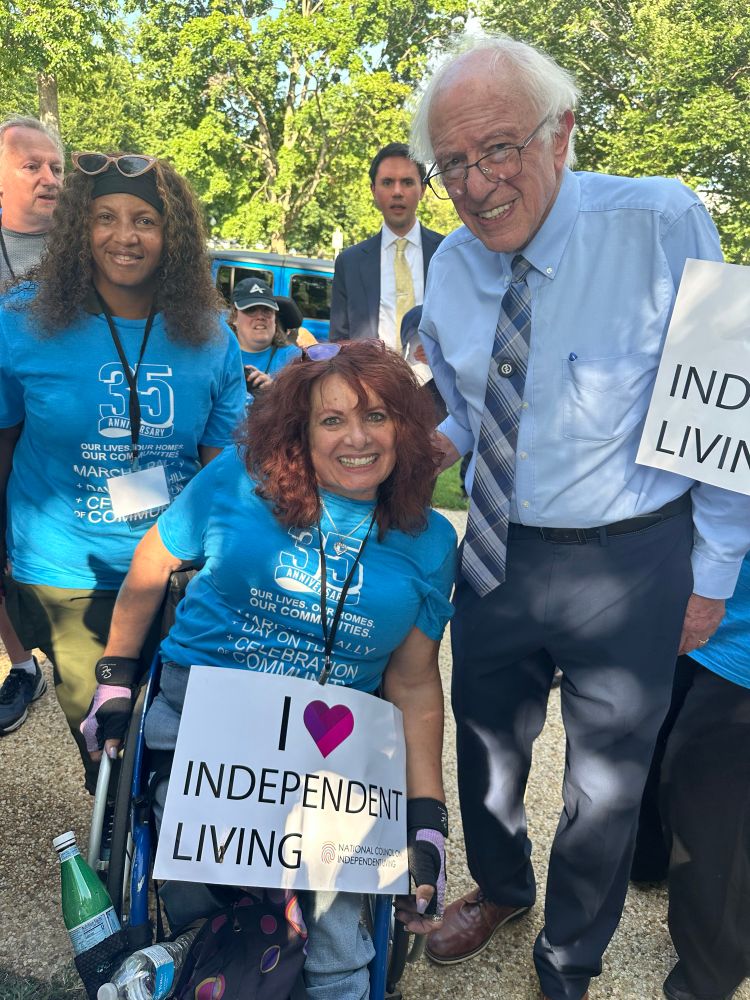 A photo of Rolling Start Inc. Executive Director, Lisa Hayes, a wheelchair user with long red hair and Program Manager, Joann Wilkes, an African American woman with long curly black hair are both wearing the blue ADA 35th anniversary shirts and are posing with Vermont Senator, Bernie Sanders, for a photo at Washington DC.