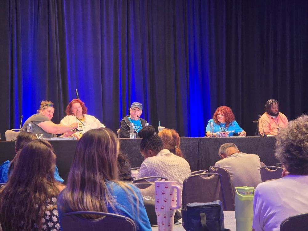 Lisa Hayes speaks on behalf of Region 9 at the NCIL Conference, seated on a panel stage alongside fellow representatives, with blue-lit curtains behind them.
