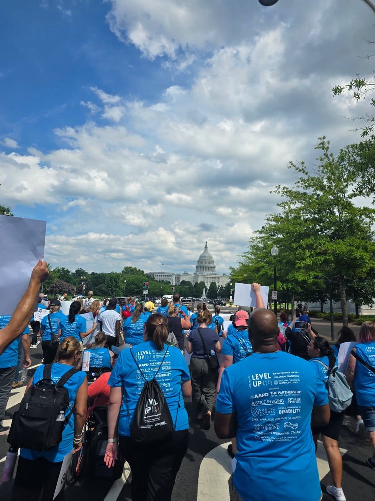 The crowd of support are marching towards the capitol building. All wearing blue 35th anniversary ADA shirts. The landscape shows the building in the distant background as the march gets closer.