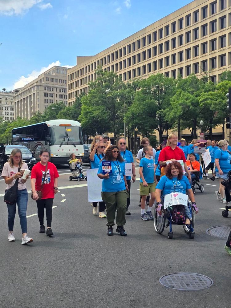 A frontal photo of the crowd, All wearing blue 35th anniversary ADA shirts on the street. Our executive director and program manager are in view, looking towards the camera as they march.