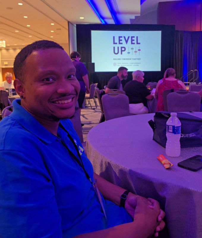 Rolling Start Inc advocate, Eddie McCreight, is sitting at a table in the audience while the panel is speaking. He is smiling at the camera. In the background a projector screen with the NCIL conference Level Up logo on screen.