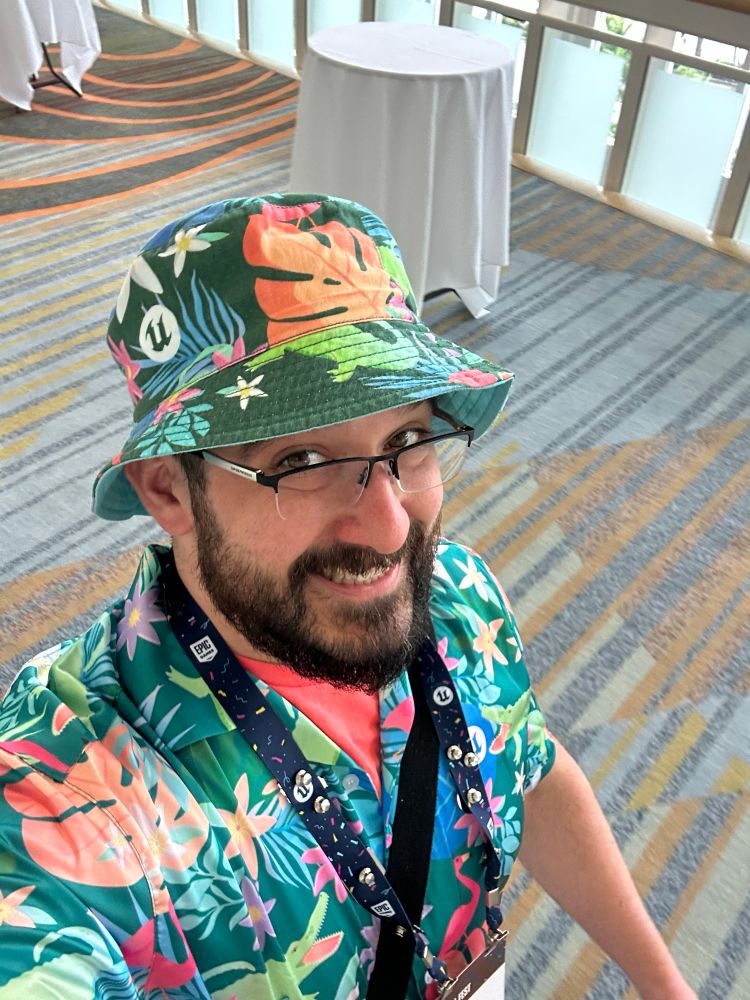 A goofy, bearded gentleman is bedecked in a matching floral hat and Hawaiian shirt. He’s cheesing for the camera. His neck is adorned by a conference badge. He is backdropped by an audacious convention center carpet pattern.