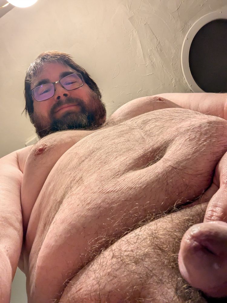 A picture from the perspective of someone on their knees looking up at a naked chubby man standing in front of them with an erection.