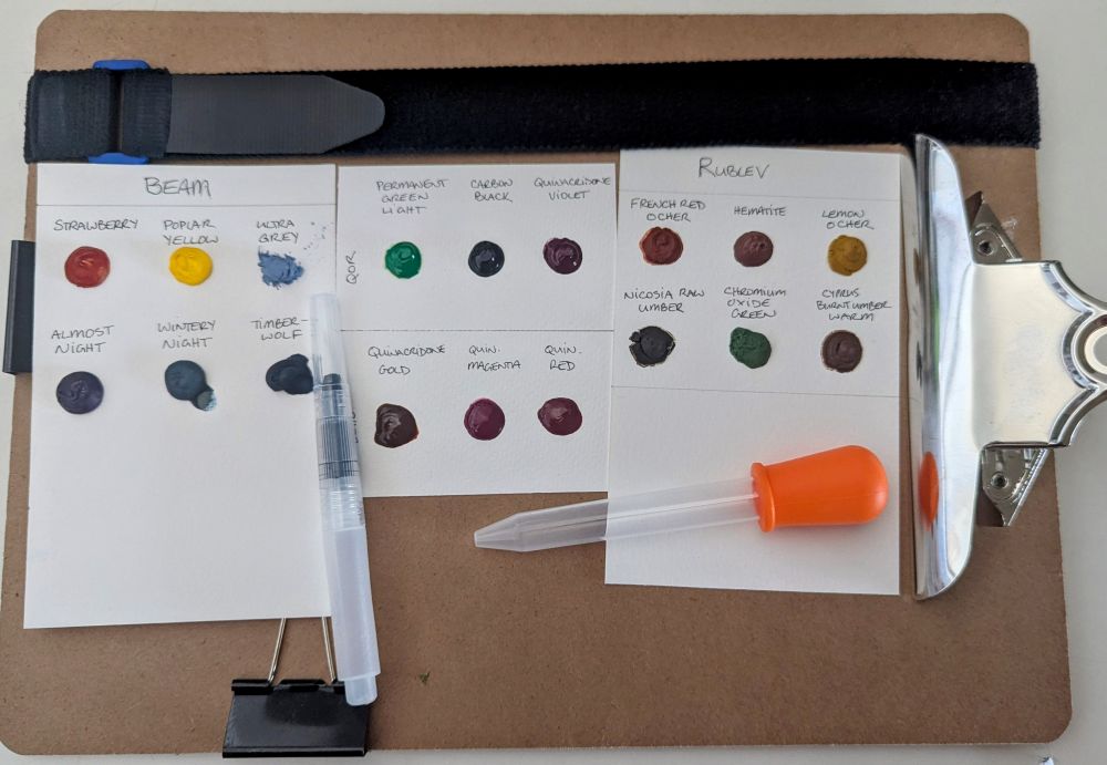 A brown clipboard laying horizontally with three cards that have round bumps of watercolour paint in them, along with a water brush and pipette. There are also clips to hold paper in place and a strap to help reduce fatigue while holding the clipboard.
