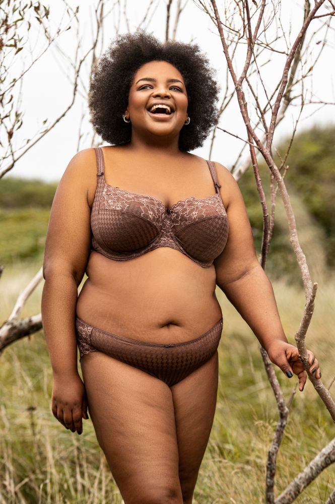 Eine Schwarze Person trägt ein dunkelbraunes Wäscheset aus Spitze und mit Hahnentrittmuster. Der BH hat Bügel und die Träger sitzen weit außen. 