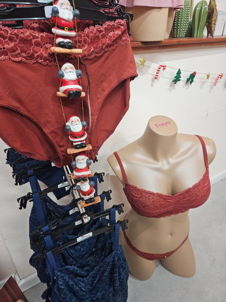 Ein roter Slip mit Spitze, darunter ein blaues Bralette hängen an einem Wandabhänger. Vor dem roten Slip hängt eine Leiter mit kleinen Weihnachtsmännern.Daneben steht eine Puppe, die ein rotes Spitzenbralette und einen String trägt. Im Hintergrund eine Girlande mit Weihnachtsbäumen, Sternen und Zuckerstangen.