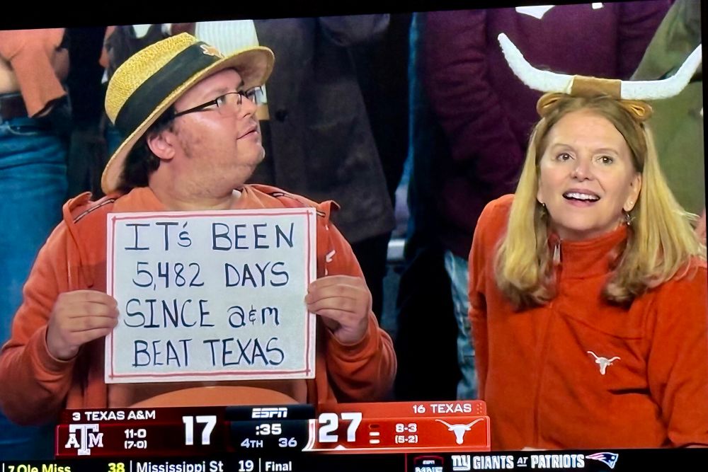 Texas fans - blonde woman wearing longhorn horns headgear while a man in a hat holds a sign saying “it’s been 5,482 days since a&m beat Texas”