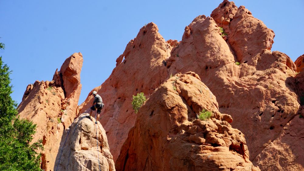 A male climber holding onto a rope leans backward at the top of a small peak with multiple rust colored stone peaks behind him 