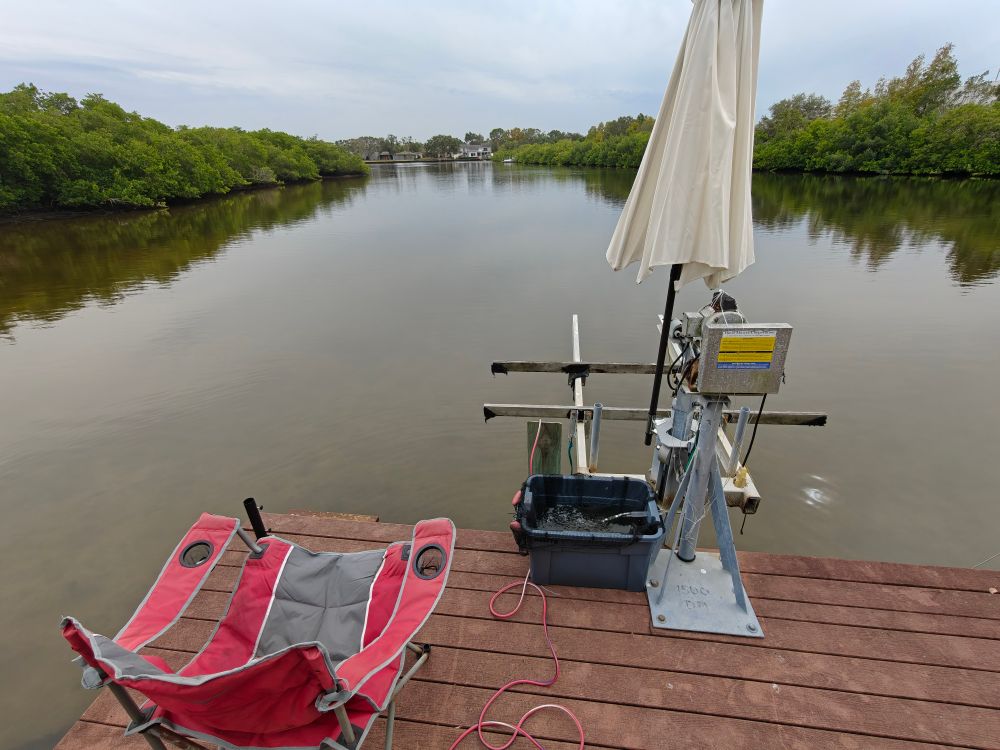 A deck chair sits on a dock overlooking water, next to a livewell and umbrella. 