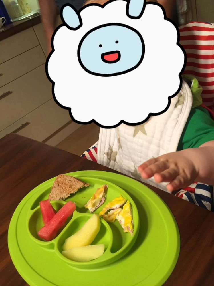 Grüner Teller aus Silikon mit 3 Unterteilungen. Gefüllt mit Wassermelone, Apfel, Toastbrot, Ei&Schinken. Eine Kinderhand greift zum Teller.