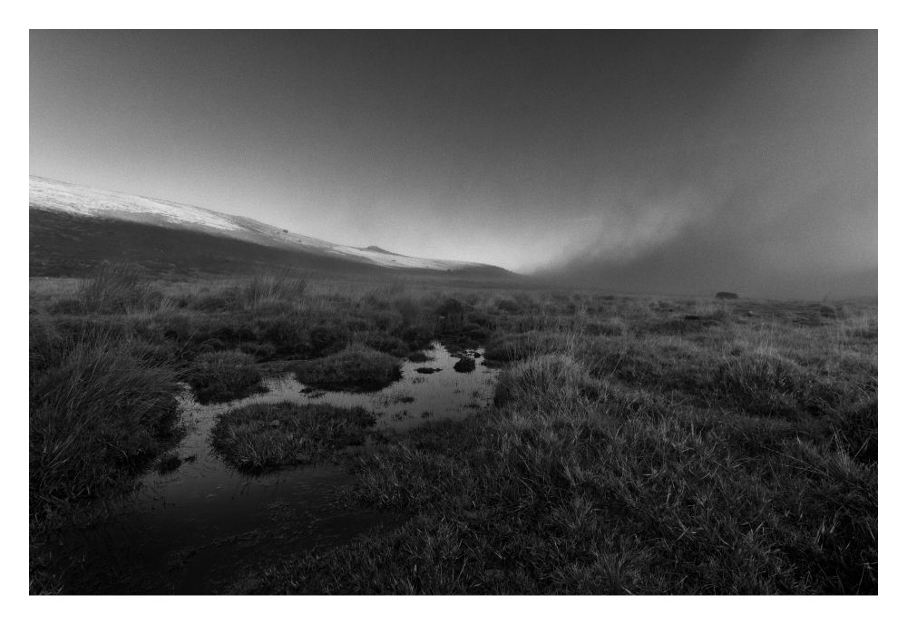 This is black and white image of a rural location in Dartmoor, in the south of the UK. In the distance you can see the light catching the tops of the hills, and on the right the low-lying fog drifting along the landscape. In the foreground is mostly wild grass, with bodies of water punctuated here and there.