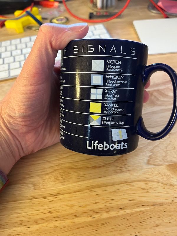 A hand holds a heavily-faded, battered charity mug from the RNLI. The last few flags of the International Code of Signals are visible: 

VICTOR: I Require Assistance
WHISKEY: I Need Medical Assistance
X-RAY: Stop Your Intention
YANKEE: I Am Dragging My Anchor
ZULU: I Require A Tug