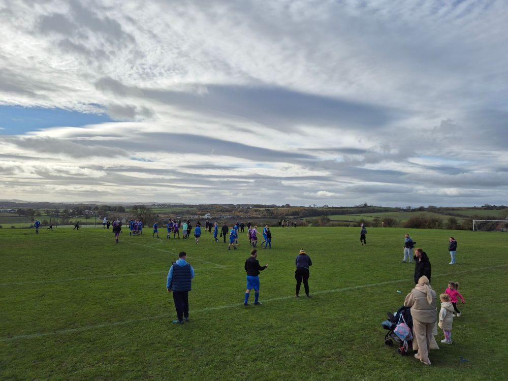 A village playing field on which two football teams have just finished their match. The background shows overcast skies, and rolling fields heading off into the distance.