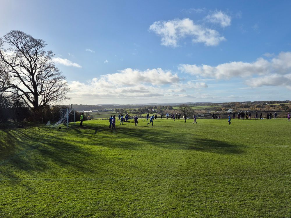 A village playing field on which two football teams are battling for the ball. The background shows blue skies, a few white clouds and rolling fields heading off into the distance.