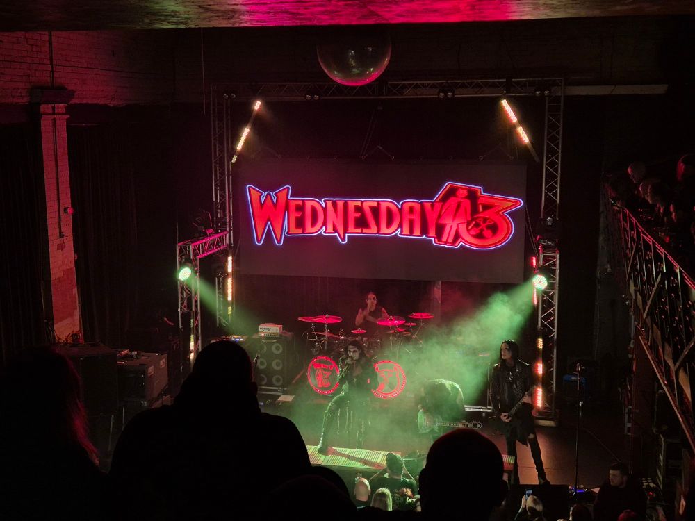 Shot of a stage taken from a balcony. A large, red Wednesday 13 logo can be seen as the backdrop, while smoky lighting spotlights the lead singer who is using a powerful rock stance. 