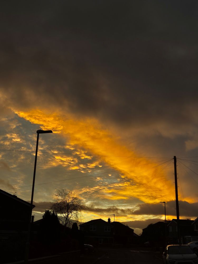 A photo of the evening sky. The top third of the picture features dark grey clouds. The bottom third shows silhouettes of the houses, trees and lampposts on the street. The middle third shows a fiery streak as the setting sun catches the clouds.