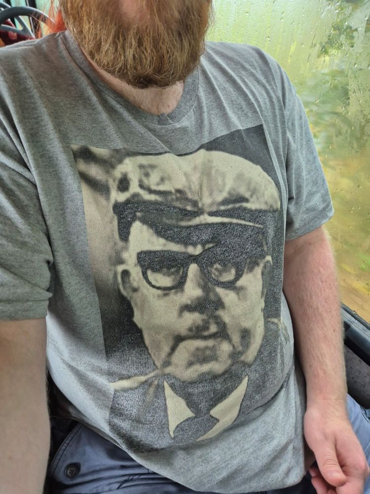 A selfie showing me sitting on the bus wearing a t-shirt that displays the face of former Coronation Street character Albert Tatlock.