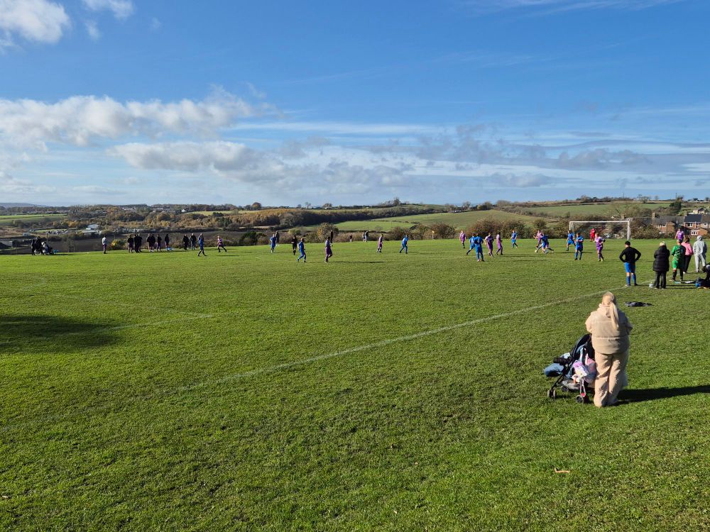A village playing field on which two football teams are battling for the ball. The background shows blue skies, a few white clouds and rolling fields heading off into the distance.