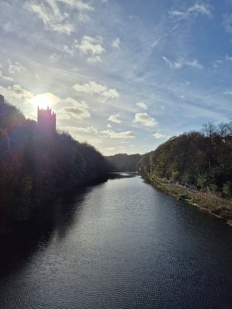 A view of the River Wear in Durham taken from Framwellgate Bridge. The sky is blue and mottled with light clouds, the sun is seen above the towers of Durham Cathedral to the left. Both sides of the river are lined with large trees.