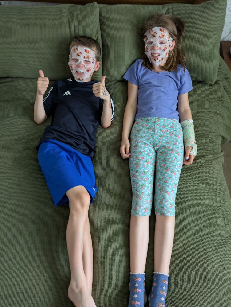 Two big kids in shorts and t-shirts lying on a bed wearing sheet face masks. The little boy on the left is putting his thumbs up. The little girl on the right has a cast on her arm.