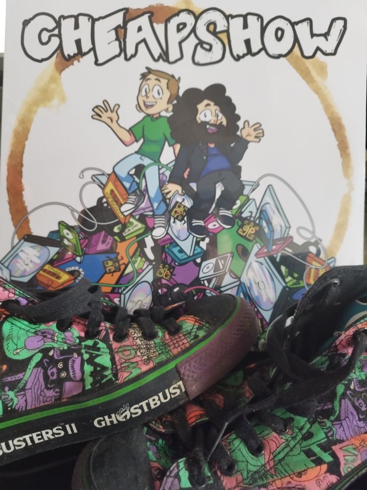 The cheapshow podcast's new album. It has 'cheaphow' at the top with hosts Paul & Eli sitting on a mountain of unwanted items that commonly are found in a charity shop. Holding the record upright are a pair of Ghostbusters II converse-style shoes.