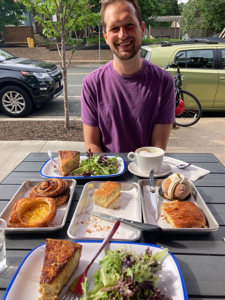A man in a purple shirt sits in front of a table filled with a variety of pastries and a cup of coffee. 