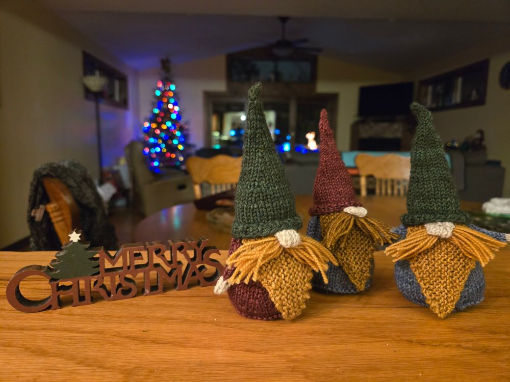 Three green, blue, and red knit gnomes with orange beards stand next to a sign saying Merry Christmas 
