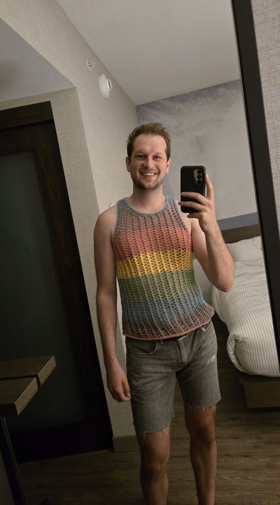 A man wearing a mesh knit rainbow tank top and jean shorts. 