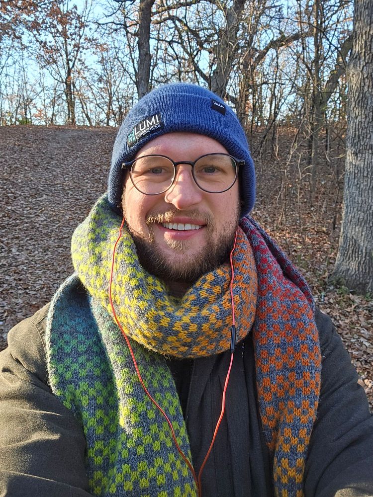 A man takes a selfie in the forest wearing a blue hat and rainbow knit scarf. 
