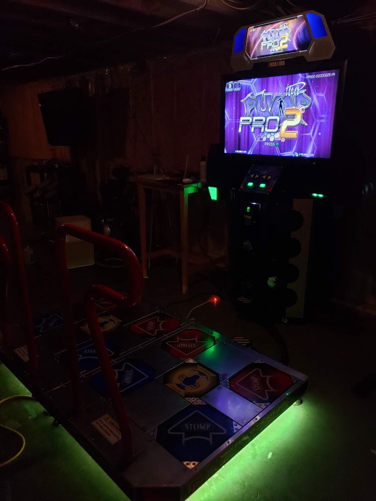 A poorly lit photo of the entire Pump it Up arcade cabinet. There's a yellow-green underglow from the RGG lights I installed underneath the stages. The arrow panels are all placed with the arrow graphics pointed inwards towards the center, instead of outwards, to reduce and even out years worth of wear on the polycarbonate during play.