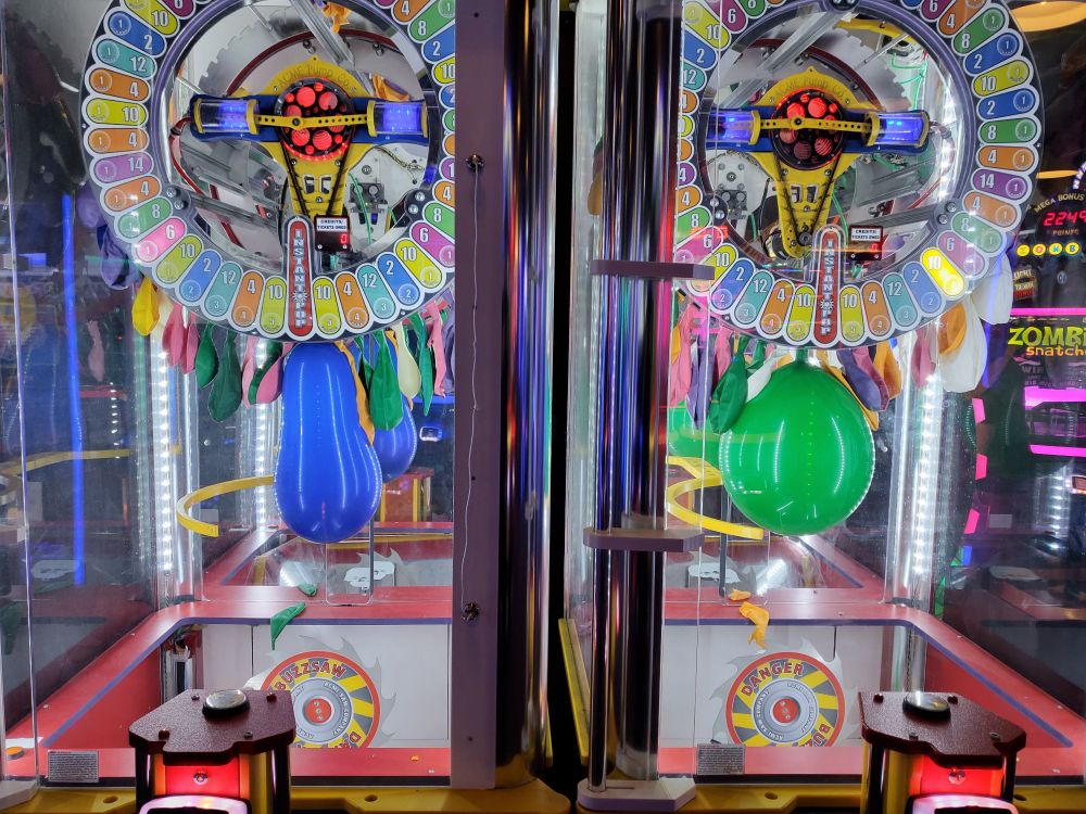 A photo of two "Explosive Danger" arcade games set up side by side. One of them has a narrow, skinny balloon. One of them has a very rotund balloon. The shapes of the balloons are oddly similar to those of the robots playable in the Portal 2 cooperative campaign.