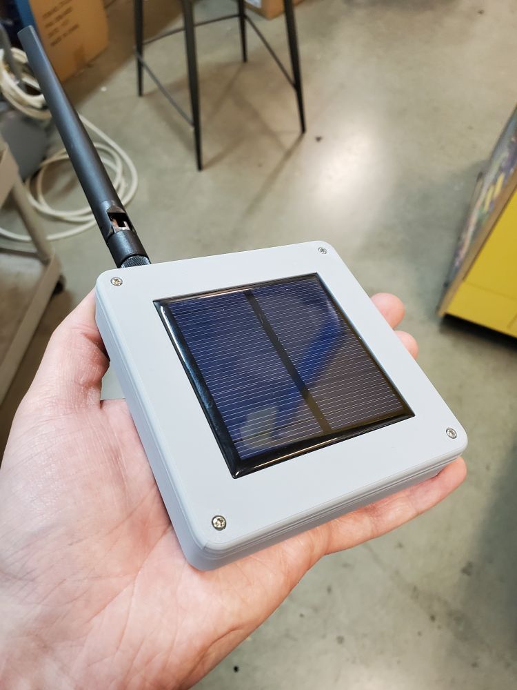 A Meshtastic node resembling a grey plastic square with screws in 4 corners, and a solar panel in the middle. There's an antenna sticking out of the upper left corner.