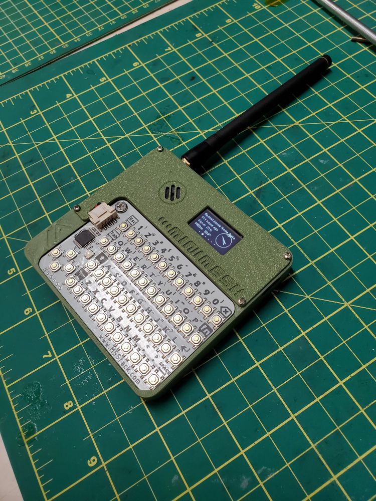 A real-life photo of the assembled Minimesh sitting on a cutting mat atop a desk. The case is printed in an olive green. The display shows information about a node titled "5panel.dance Home Base". There is a long antenna attached to the upper left corner.