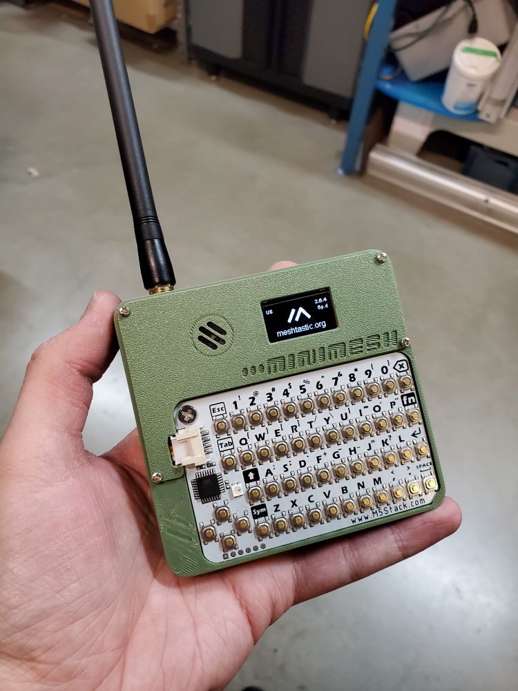A real-life photo of the assembled Minimesh, being held in my left hand. The case is printed in an olive green. The display shows the Meshtastic logo, and some miscellaneous metadata. There is a long antenna attached to the upper left corner.
