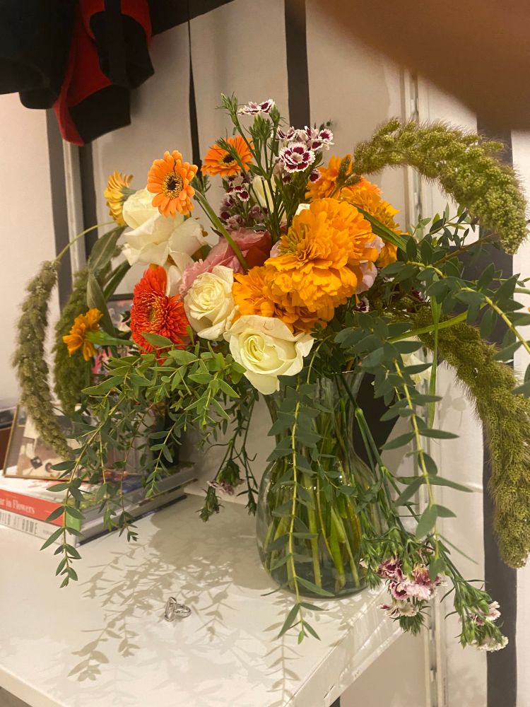 The same vase filled with a vibrant bouquet of orange, yellow, and red flowers and long, leafy green stems sits on a white shelf from another angle. Books & a picture frame are seen in background 