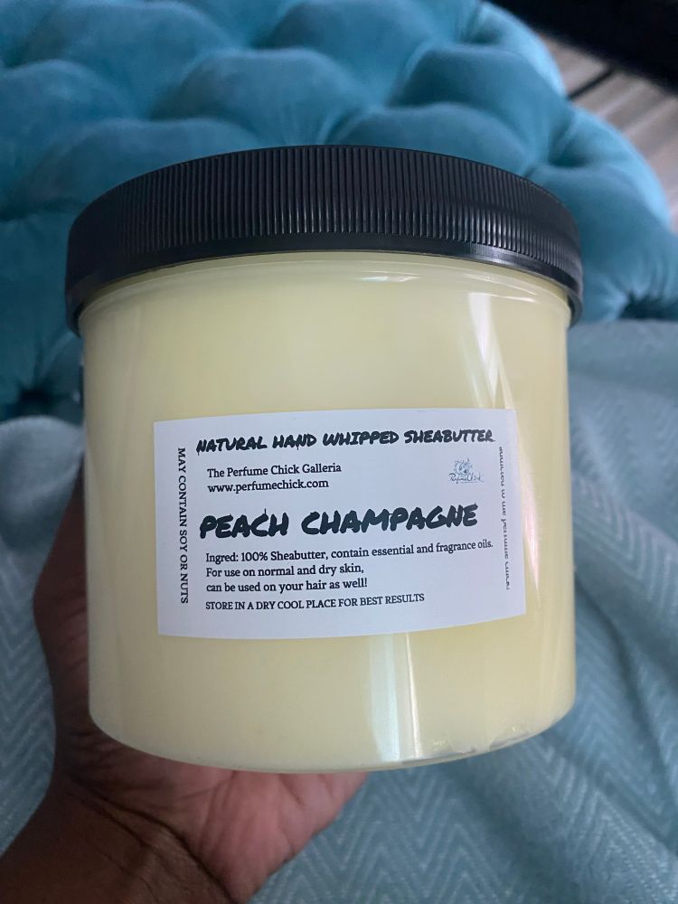 A large, clear plastic jar with a black lid, filled with a light yellow, creamy shea butter. A white label on the front reads "NATURAL HAND WHIPPED SHEABUTTER" at the top, followed by "The Perfume Chick Galleria" and "www.perfumechick.com". Below that, in larger, decorative script, it says "PEACH CHAMPAGNE". Further down, smaller text reads: "Ingred: 100% Sheabutter, contain essential and fragrance oils. For use on normal and dry skin. Can be used on your hair as well! STORE IN A DRY COOL PLACE FOR BEST RESULTS". A warning on the left side of the label says "MAY CONTAIN SOY OR NUTS". @terrysburner is holding the jar m and a light blue, blankey is visible in the background.