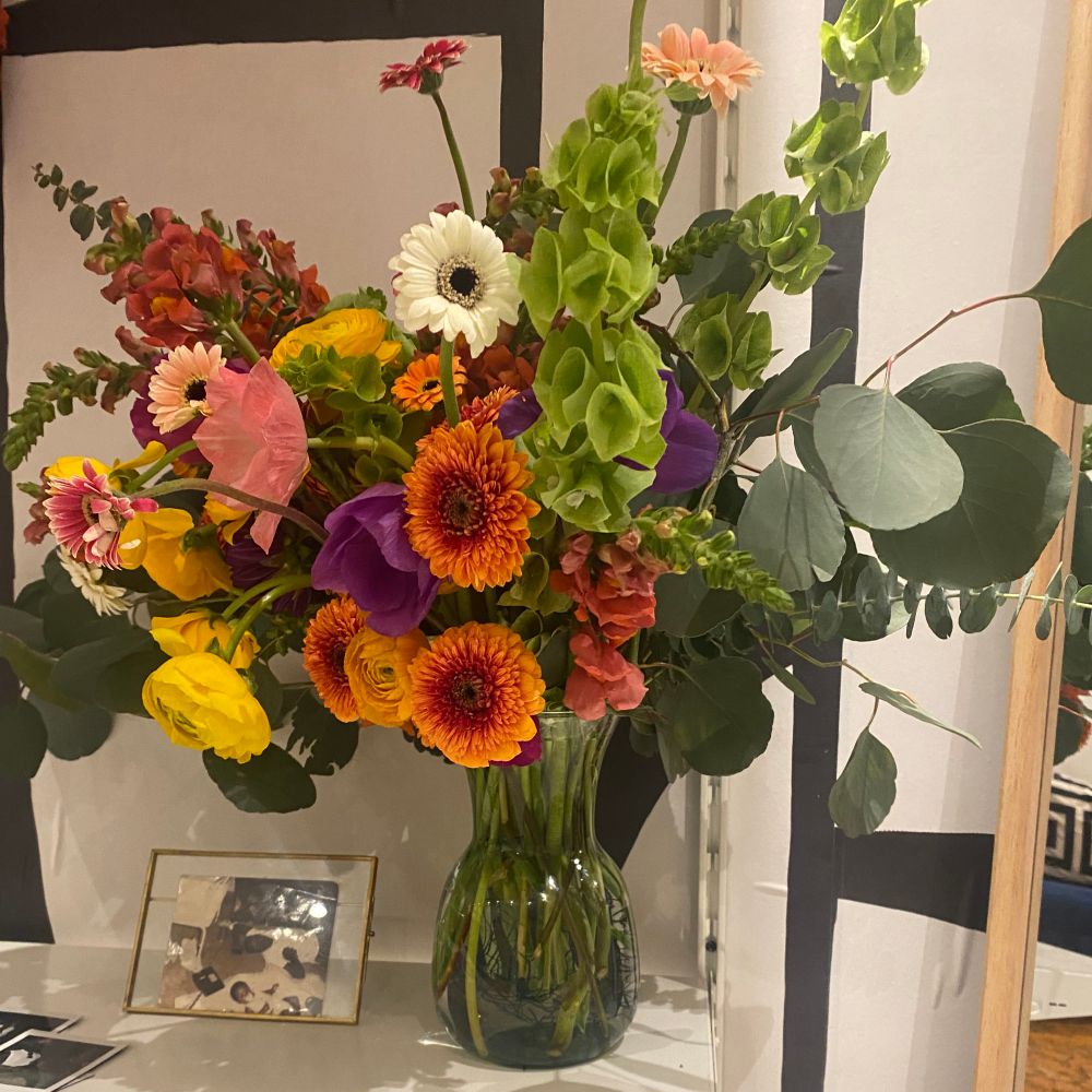 A tall glass vase holds a large, vibrant bouquet of various flowers including gerbera daisies, ranunculus, white flowers, and green bells of Ireland, with eucalyptus leaves. The vase sits on a white shelf next to a small framed photo.