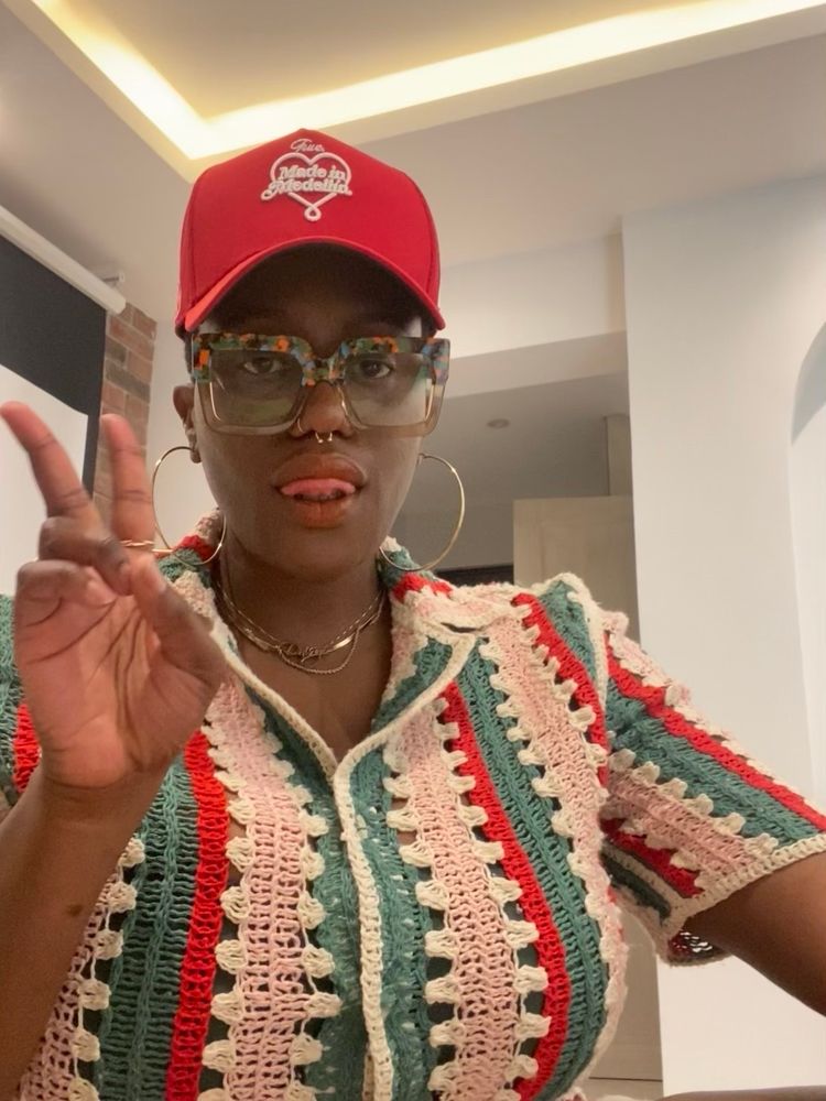 Me in a colorful crochet top & red hat holding ip a peace sign 