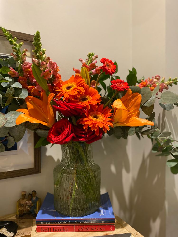  A floral arrangement in a textured clear glass vase sits on top of a small stack of books. The bouquet contains a variety of colorful flowers, including orange lilies, orange gerbera daisies, red roses, and pink snapdragons. The arrangement is accented with long, trailing eucalyptus branches.