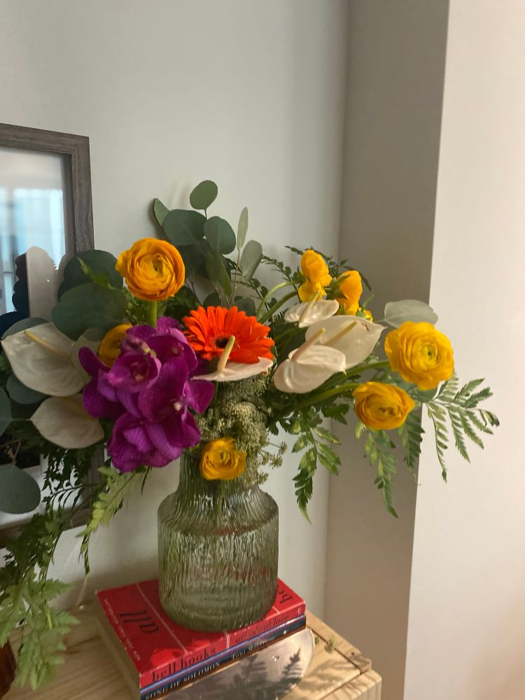 A close-up of a floral bouquet in a textured glass vase, placed on red and blue stacked books. The bouquet includes bright yellow ranunculus, a vivid purple orchid, white anthuriums, and an orange gerbera daisy, accented with green foliage.