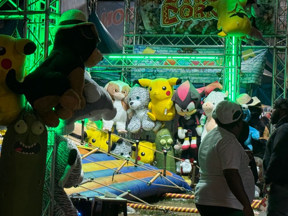 A gloomy shadow the hedgehog plush strung up between pikachu, pickle rick, and a cow of sorts. The neon green glow of the silly rope game setup at night illuminates their cheap polyester heads